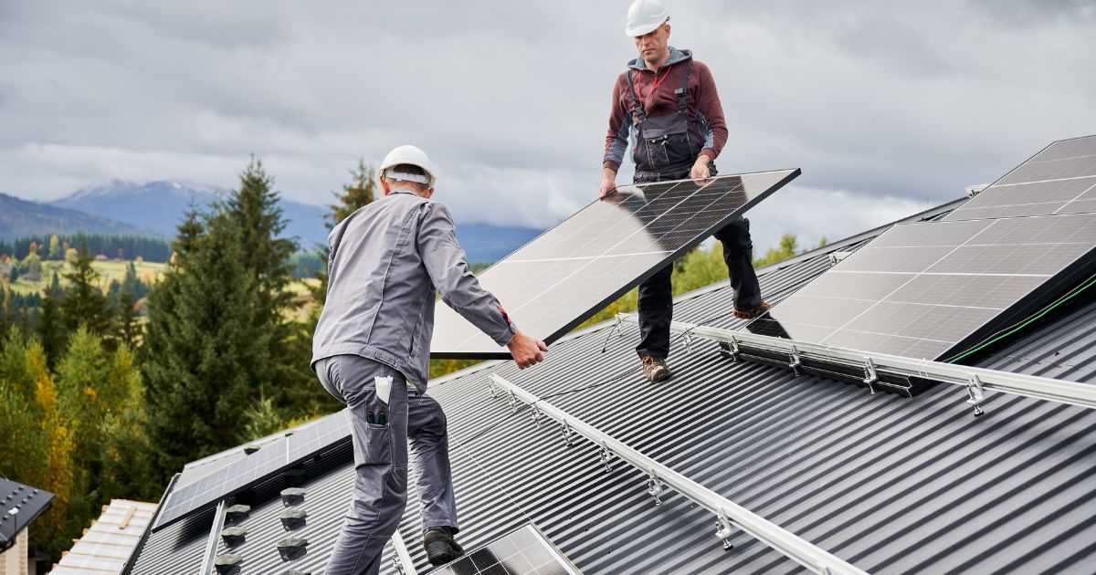 Doi instalatori montează panouri solare pe acoperișul unei case, cu munții Carpați în fundal - exact cum arată o comunitate de energie în timpul implementării